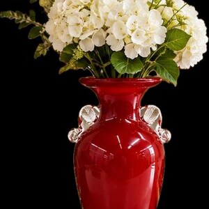 May include: A glossy red glass vase with two clear, leaf-shaped handles on the sides. The vase is filled with a large, dense bouquet of white hydrangea flowers and green leaves, creating a high-contrast display against the dark background.