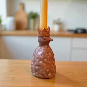 Könnte beinhalten: Ein handgefertigter Kerzenhalter aus Keramik in Form eines Vogels, gekrönt mit einer kleinen Krone. Der vogelformige Halter ist braun mit einer strukturierten Oberfläche und hält eine hohe, gelbe Kerze. Der Kerzenhalter steht auf einem Holztisch.
