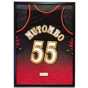 Può includere: Maglia da basket nera incorniciata con il nome "MUTOMBO" e il numero "55" in bianco e oro. La maglia presenta dettagli rossi e dorati ed è presentata in una cornice nera.