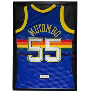Può includere: Maglia da basket blu incorniciata con finiture gialle, il nome "MUTOMBO" e il numero "55" in bianco. Una striscia arcobaleno attraversa il petto. La maglia è esposta in una cornice nera.