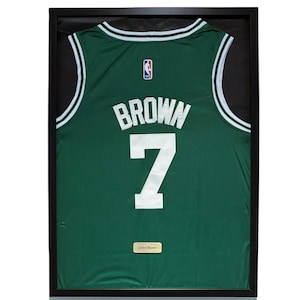 Puede incluir: Camiseta de baloncesto verde enmarcada con detalles blancos, con el nombre "BROWN" y el número "7" en blanco. El logotipo de la NBA es visible. Una pequeña placa en la parte inferior dice "Jaylen Brown". La camiseta está en un marco negro.