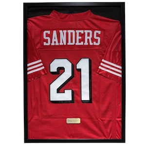Pode incluir: Camisola de futebol vermelha emoldurada com o nome "SANDERS" nas costas e o número "21" em branco. A camisola tem listras brancas nas mangas e é exibida em uma moldura preta.