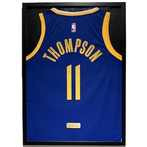 Peut inclure: Maillot de basket-ball bleu encadré avec des bordures dorées, le nom "THOMPSON" et le numéro "11" en or. Le maillot porte le logo NBA et est présenté dans un cadre noir.