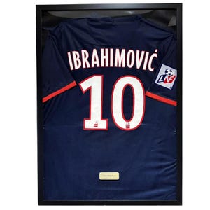 Pode incluir: Camisola de futebol azul marinho emoldurada com o nome "IBRAHIMOVIC" e o número "10" em branco. A camisola tem detalhes em vermelho e um logótipo da equipa na manga. A camisola é exibida numa moldura preta.