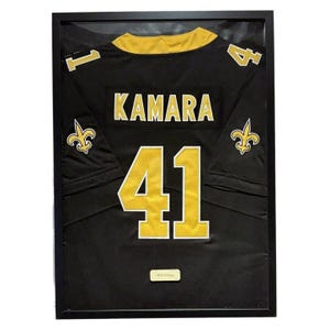 Peut inclure: Maillot de football noir et or encadré. Le maillot porte le nom "KAMARA" et le numéro "41" en or. Le maillot porte également les numéros "1" et "4" sur les épaules. Le maillot est encadré dans un cadre noir.