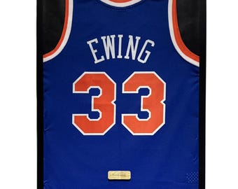 May include: Framed blue basketball jersey with the name "EWING" and the number "33" in white and orange. The jersey has white and orange trim around the collar and armholes. The jersey is displayed in a black frame.