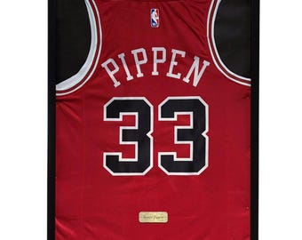 May include: Framed red basketball jersey with white and black detailing. The jersey displays the name "PIPPEN" and the number "33" in white with black outlines. A small wooden plaque with "Scottie Pippen" is at the bottom.
