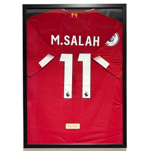 May include: Framed red soccer jersey with the name "M. SALAH" and the number "11" in white. The jersey is displayed in a black frame, showcasing the team's logo and a Premier League emblem. A small gold nameplate is at the bottom.