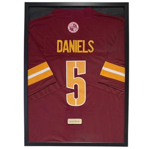 Pode incluir: Camisola de futebol bordô emoldurada com detalhes dourados e brancos. A camisola exibe o nome "DANIELS" e o número "5" em dourado. As mangas têm listras douradas e brancas. Uma pequena placa na parte inferior diz "Jayden Daniels".