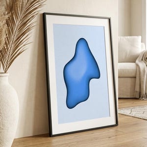 Peut inclure: Impression d'art encadrée abstraite avec une forme bleue organique sur fond bleu clair. L'œuvre est dans un cadre noir et est appuyée contre un mur. Un vase blanc avec des plantes séchées est au premier plan.