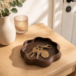 Könnte beinhalten: Ein braunes, blütenförmiges Tablett hält Schlüssel mit einem goldfarbenen Schlüsselanhänger mit der Aufschrift "Home Sweet Home". Das Tablett steht auf einem Holztisch neben einer Vase mit Blumen und einem Kerzenhalter.