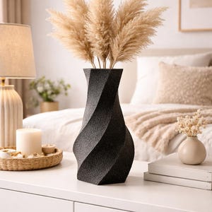 Könnte beinhalten: Eine schwarze, strukturierte Vase mit gedrehtem Design hält einen Bund beiger Pampasgras. Die Vase steht auf einer weißen Oberfläche, mit einer Kerze und einer Lampe im Hintergrund. Die Vase ist etwa 30 cm hoch.