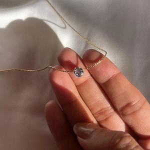 Könnte beinhalten: Eine zarte goldene Halskette mit einem einzelnen, klaren, runden Edelsteinanhänger. Die Halskette wird vor einem weichen, weißen Hintergrund gehalten, der die schlichte Eleganz des Schmucks hervorhebt. Die Kette ist dünn und der Edelstein funkelt.