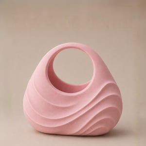 May include: A light pink ceramic vase with a handle. The vase has a rounded shape with a series of wave-like ridges on the front. The handle is a large, open circle. The vase is a solid, soft pink color.