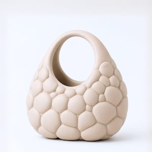 Op de afbeelding: Een beige handtas met een bubbeltextuur en een ronde handgreep. De tas heeft een druppelvorm en is bedekt met afgeronde, onregelmatige vormen. Het unieke ontwerp en de neutrale kleur maken het een opvallend accessoire.
