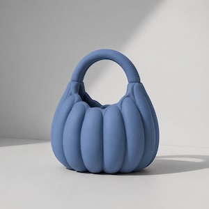 Op de afbeelding: Een matte blauwe handtas met een ronde, pompoenachtige vorm met verticale geribbelde segmenten en een enkel geïntegreerd boogvormig handvat. De tas heeft een gladde, uniforme textuur en een effen kleur over de gehele gebogen structuur.