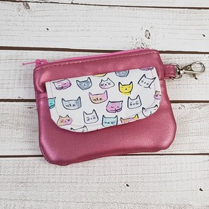 May include: A pink zippered pouch with a white fabric lining featuring a pattern of colorful cat faces. The pouch has a metal clasp and a small zippered pocket on the front.