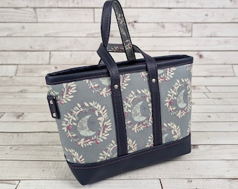 May include: A blue and gray tote bag with a floral pattern and a crescent moon design. The bag has a dark blue leather trim and two top handles.