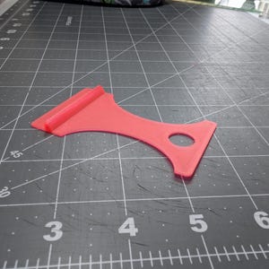 May include: A coral-colored plastic tool with a unique shape, featuring a rounded handle with a hole and a rectangular blade. The tool is designed for a specific purpose, likely for crafting or DIY projects.