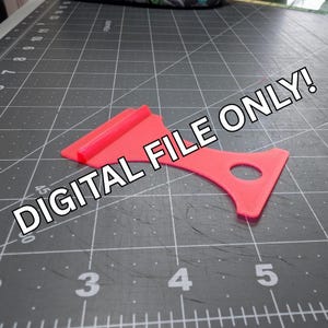 Könnte beinhalten: Ein leuchtend rosa Kunststoffwerkzeug mit einer flachen Kante und einem gebogenen Griff. Das Werkzeug hat ein kleines Loch in der Nähe des Griffs und den Text "DIGITAL FILE ONLY!". Es liegt auf einer grauen Schneidematte.