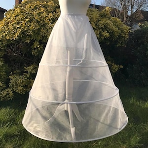 May include: A white bridal petticoat with three hoops is displayed on a mannequin. The petticoat is made of sheer fabric and has a full, A-line shape. A small floral headpiece adorns the mannequin's head. The background features greenery and a clear sky.