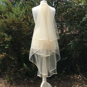 May include: A bridal veil on a white mannequin. The veil is a soft, off-white color and has a layered design with a delicate trim. The mannequin is on a white stand, set against a backdrop of green foliage and purple flowers.