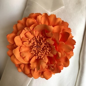 Könnte beinhalten: Eine leuchtend orangefarbene Blumen-Boutonniere mit geschichteten Blütenblättern und einem strukturierten Zentrum. Das florale Accessoire ist an einem weißen Blazer befestigt und erzeugt einen auffälligen Kontrast. Dieses dekorative Stück setzt einen Farbtupfer.