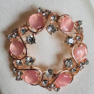 Puede incluir: Broche de color oro rosa con un diseño circular. La broche presenta piedras rosas ovaladas y cristales transparentes. Las piedras están engastadas en un delicado patrón arremolinado, creando un accesorio elegante.