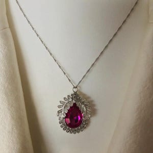 May include: A silver necklace with a large, fuchsia-coloured, teardrop-shaped pendant. The pendant is set in an ornate, openwork frame with clear accent stones, suspended from a delicate chain.