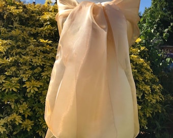 May include: A sheer, golden-toned shawl draped over a mannequin. The fabric is gathered and tied at the front, creating a layered, flowing effect. The background features lush green foliage and a bright blue sky.