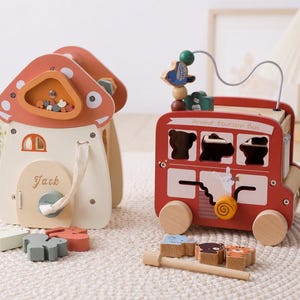 May include: A wooden mushroom-shaped activity house with a door labeled Jack and a red wooden bus toy labeled Animal Tourism Bus. Both toys feature various cutouts, sliding elements, and small wooden animal-shaped pieces for sorting activities.