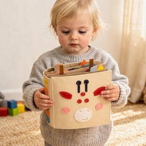 May include: A beige fabric activity book with a giraffe face design. The book has colorful shapes and tabs. A child holds the book, with a stuffed bunny, teddy bear, and building blocks in the background.