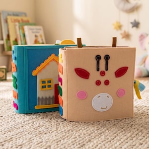 May include: Two colorful felt activity books for toddlers. One is blue with a house design, the other is tan with a giraffe face. Other toys and books are in the background, creating a playful scene.