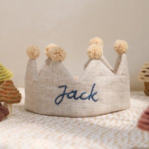 May include: A beige fabric crown with the name "Jack" embroidered in blue script. The crown has six beige pom-poms on top. Decorative mushroom and flower-shaped items surround the crown.