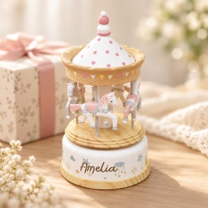 May include: A wooden carousel music box featuring white and pink horses, a white roof with pink polka dots, and a base printed with the name Amelia, surrounded by small star and cloud illustrations in soft pastel colors and natural wood tones.