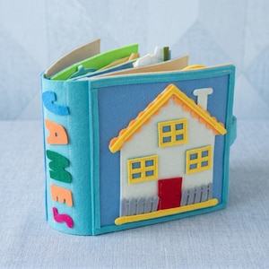 Peut inclure: Un livre calme en feutre fait à la main avec une couverture bleue portant le nom "JAMES" en lettres colorées. La couverture avant présente une maison avec un toit jaune, des murs blancs et une porte rouge. Les pages du livre sont visibles.