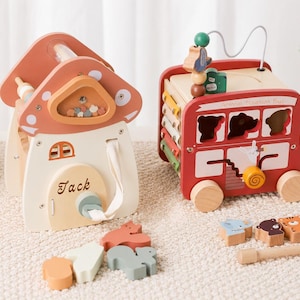 May include: A wooden mushroom-shaped activity house with the name Jack printed on the door and a red wooden bus-shaped activity toy labeled Animal Tourism Bus, both featuring various interactive elements, sliding parts, and small animal-shaped blocks.