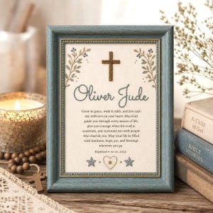 May include: A framed baptismal keepsake featuring a blue wooden frame, a central embroidered cross, and the name Oliver Jude. The text includes a blessing, the date 10.10.2025, and decorative embroidered floral sprigs, two stars, and a heart.