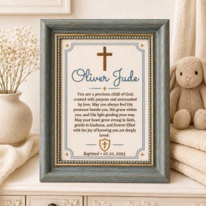May include: A rectangular, distressed blue-grey wooden frame containing a white fabric insert with a brown cross, the name Oliver Jude, a religious blessing, a small shield icon, and the text Baptized 10.10.2025 in dark blue and gold lettering.