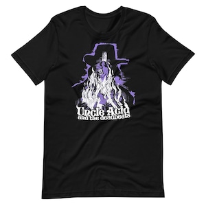 May include: Black t-shirt featuring a purple and white graphic of a figure in a hat with long hair. The text "Uncle Acid and the deadbeats" is printed below the graphic in white.