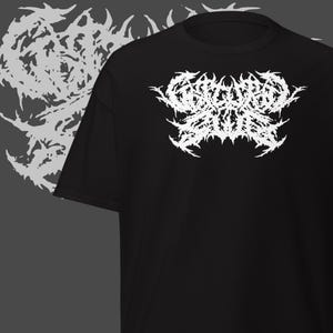 Puede incluir: Camiseta negra con un diseño gráfico blanco intrincado, posiblemente un logotipo de banda. Una versión descolorida del diseño es visible a la izquierda. La camiseta está hecha de un material suave. El diseño está centrado en la parte delantera.