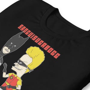 May include: Black t-shirt featuring cartoon characters in a parody of Batman and Robin. The text "SANGUISUGABOGG" is printed in red. The shirt has a crew neck and short sleeves.