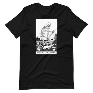 May include: Black t-shirt featuring a white graphic of a skeleton riding a bicycle, with a sunset and the text "HIGH ON FIRE". The design includes two faces and a crown. The shirt is made of a soft material.