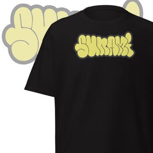 May include: Black t-shirt with the word "SURRAY" in yellow bubble letters. The shirt has a crew neck and short sleeves. A yellow hand graphic is visible in the background.