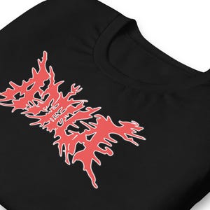 Könnte beinhalten: Schwarzes T-Shirt mit einem roten, stilisierten Grafikdesign. Das Design hat eine gezackte, organische Form mit weißen Umrissen. Das Shirt hat einen Rundhalsausschnitt und kurze Ärmel, aus weichem Material.