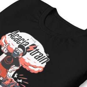 May include: Black t-shirt with a cartoon illustration of a man with glasses and a beard, giving a thumbs up. The design includes the text "The Acacia Strain" in white and red, with a lightning bolt graphic.