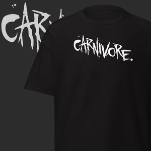 May include: Black t-shirt with the word "CARNIVORE" in white, stylized, distressed lettering. The design is repeated in a faded, mirrored effect on the left side. The shirt is a casual wear item.