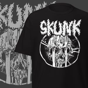 May include: Black t-shirt with a white graphic featuring the word "SKUNK" above a skull design. The skull is inside a broken circle. The design has a heavy metal or punk rock aesthetic.