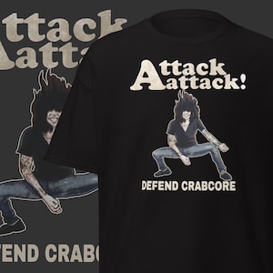 Op de afbeelding: Zwart T-shirt met de tekst "Attack Attack!" en een afbeelding van een persoon in een krabachtige pose. De tekst "DEFEND CRABCORE" staat onder de afbeelding. Een vergelijkbaar ontwerp staat op een grijze achtergrond.