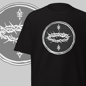 May include: Black t-shirt featuring a white graphic of a crown of thorns within a circle. The design includes a stylized cross at the top and bottom of the circle. A similar design is visible in the background on a gray circle.